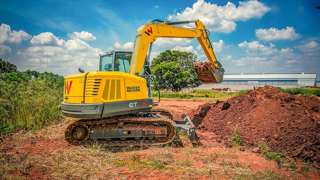 Une mini-pelle jaune de la marque Wacker Neuson, modèle ET75, soulève de la terre rouge avec sa pelle sur un chantier par temps ensoleillé. À l'arrière-plan, on aperçoit de la végétation verte, un bâtiment industriel et un ciel bleu parsemé de nuages blancs.